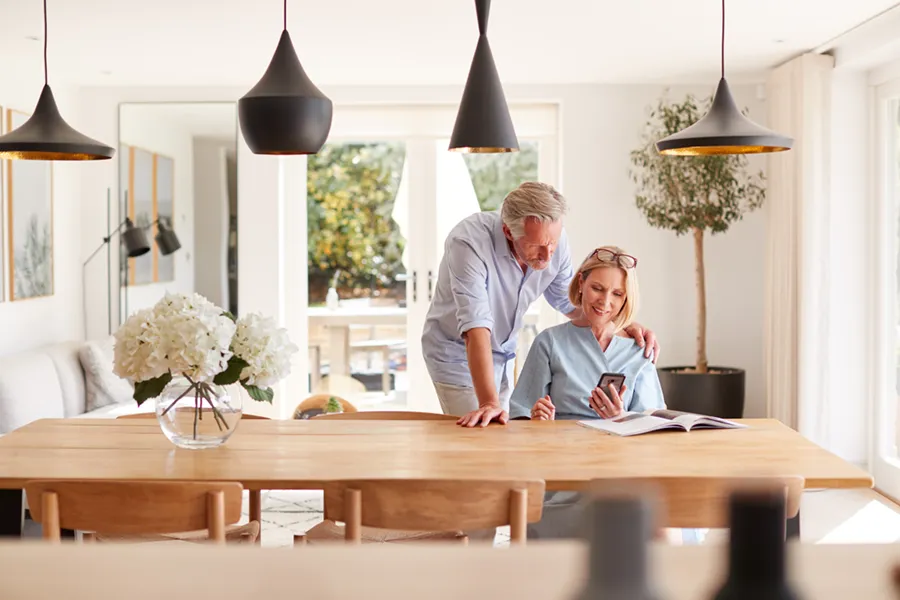 An elderly couple in their dining room, looking at something on her mobile phone.