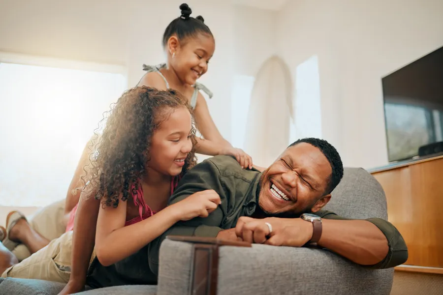 A father playing on the couch with his young daughters in their air-conditioned home.