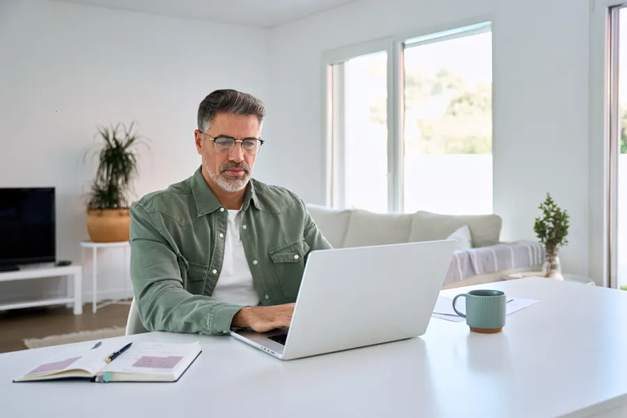 A middle-aged man siting at the table in his home and using a laptop to look up information.