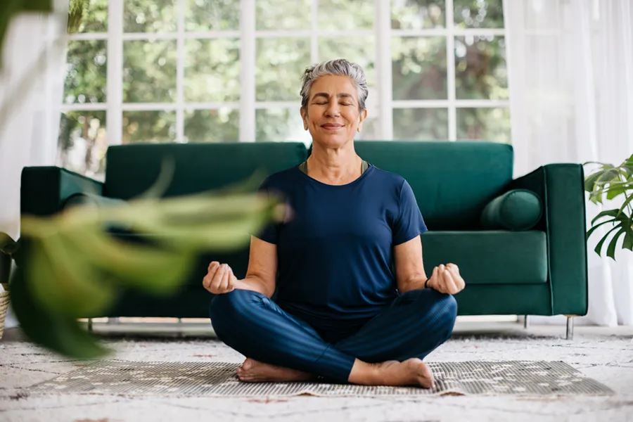 A middle-aged woman sitting cross-legged on the floor in her living room, meditating with her eyes closed.