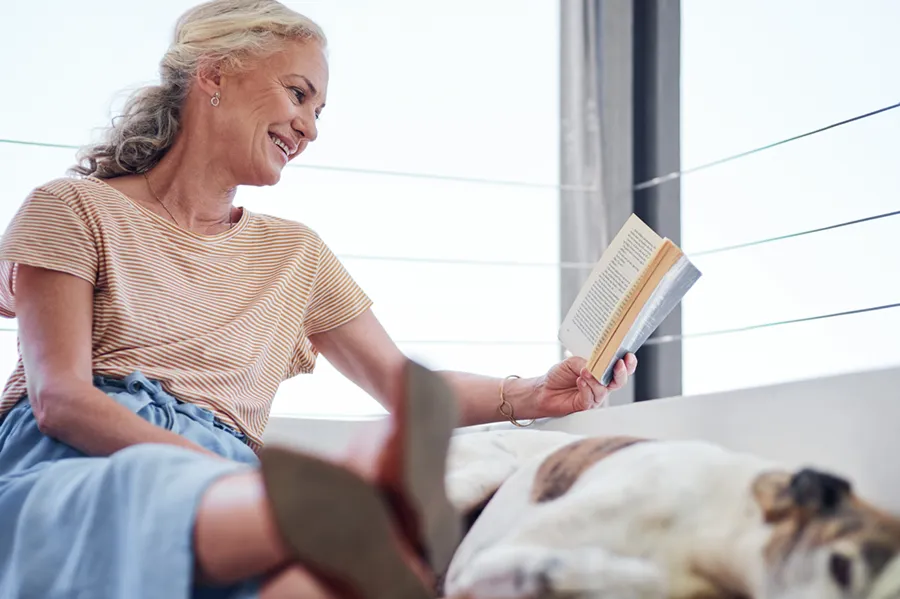 A senior woman reading a book on her couch indoors, relaxing alongside her dog.