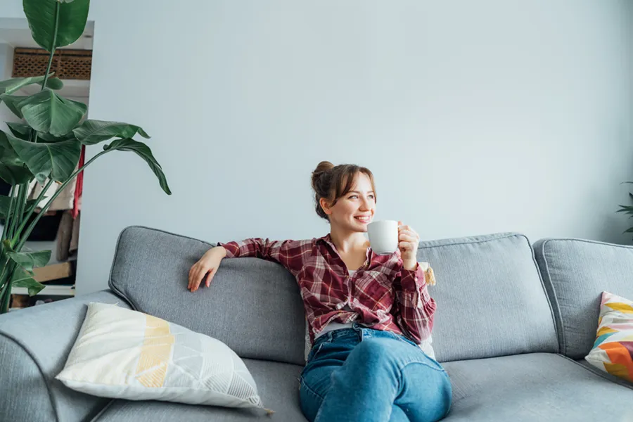 A young woman sitting on her couch and drinking a warm beverage, enjoying the central air conditioning in her home.