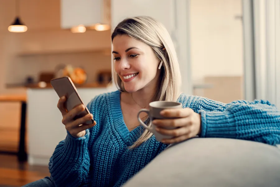 Young woman lounging on her couch with a hot beverage, looking at something on her smartphone and smiling.