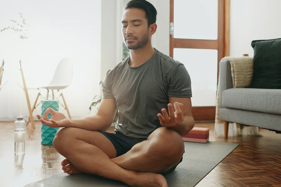 A young Asian-American man sitting cross-legged on the floor in his living room, meditating with his eyes closed.